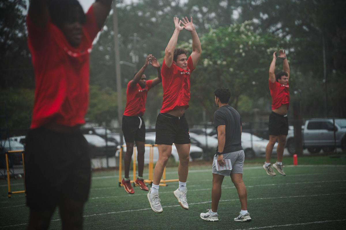 Baseball players jumping in a warm-up drill on a wet morning field while Kelvin coaches.