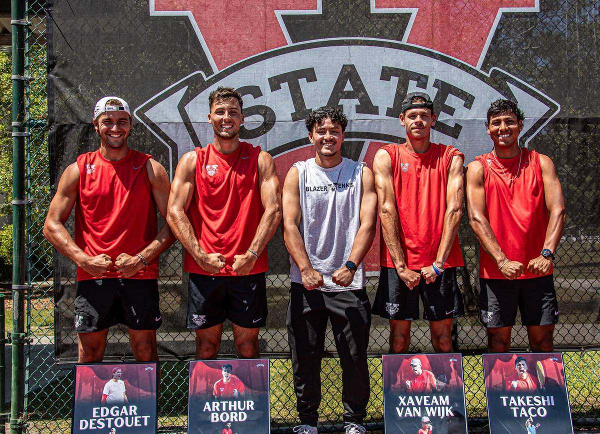 Kelvin standing with four Valdosta State men's tennis players in front of a team banner.
